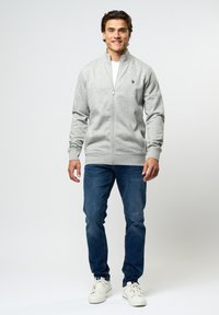 Grå hettejakke med høy krage, ribbede mansjetter og en liten logo på venstre bryst, kombinert med blå jeans og hvite sneakers.