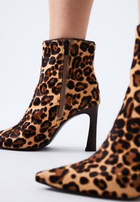 Botas de estampado de leopardo hasta el tobillo con cremallera lateral y tacones altos delgados, usadas sobre piernas desnudas contra un fondo sencillo.