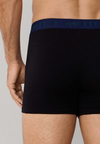 Mężczyzna w czarnych bokserkach z granatowym paskiem w pasie z napisem "HACKETT", widziany od tyłu, kadrowany powyżej talii.