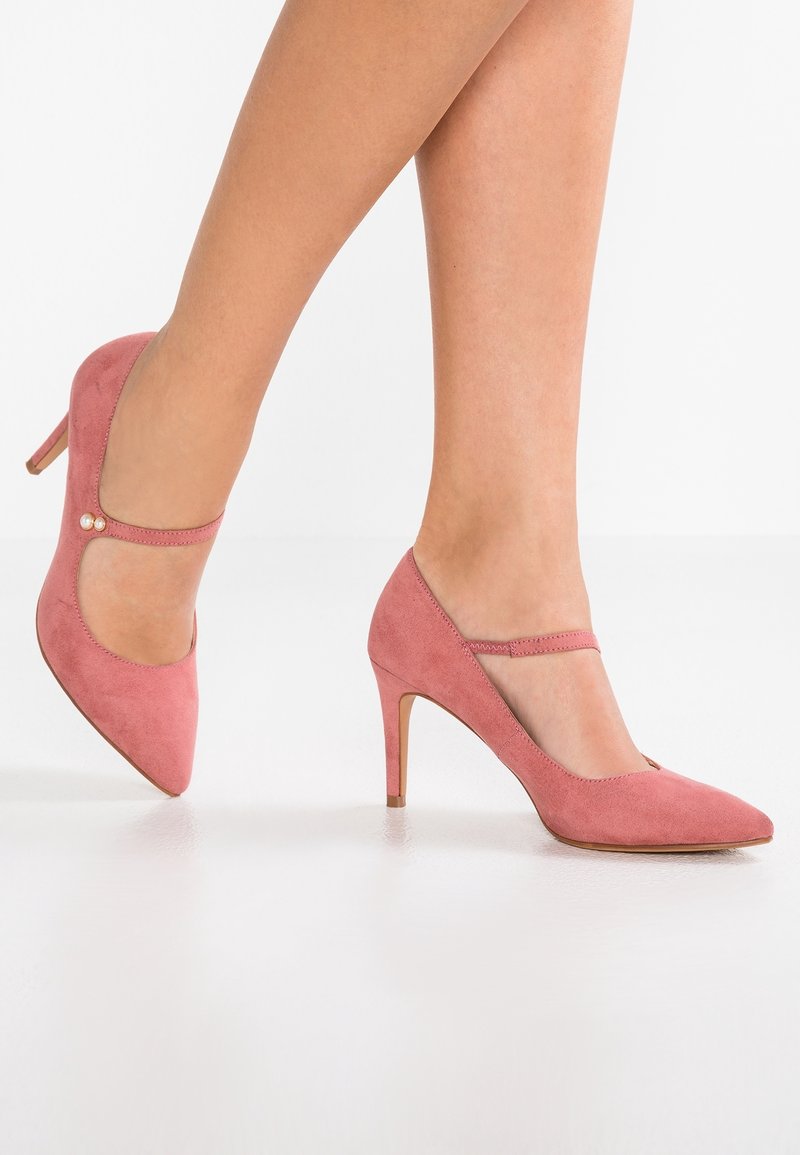 Jambes portant des talons hauts en daim rose avec des brides Mary Jane et de petits boutons en perle sur un fond blanc.