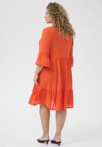 Orange, knälång klänning med en utsvängd kjol, trekvarts ärmar med klockform och en tierad rynkad fåll. Bärs med svarta muleklackar.