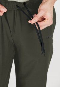 Dunkelgrüne Jogginghosen mit einem Kordelzug-Bund, ausgestattet mit schwarzen Nylon-Schnürsenkeln und einem dezenten Logo auf dem Oberschenkel. Glattes, leichtes Material.
