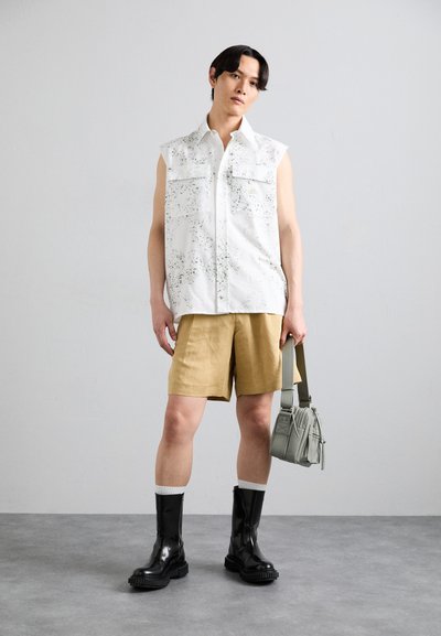 Maglietta bianca senza maniche con motivo a macchie verdi, pantaloni corti color khaki, stivali neri lucidi al ginocchio e una piccola borsa a tracolla verde chiaro.