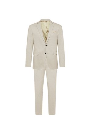 Man wearing beige suit with elastic waist trousers, white shirt, and black dress shoes against a plain white background.