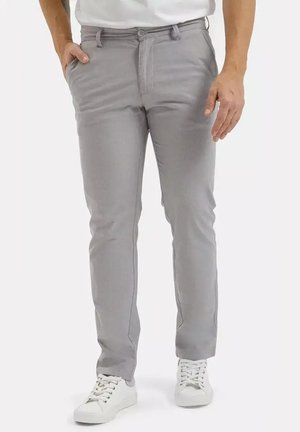 Mężczyzna w jasnoszarych spodniach typu slim fit i białych trampkach, stojący z jedną ręką w kieszeni, a drugą swobodnie opartą wzdłuż ciała.