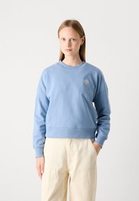 Sweatshirt bleu clair à encolure ronde et ourlet côtelé. Présente des épaules tombantes et un petit détail brodé en or. Accompagné de pantalons clairs.