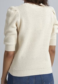 Cremefarbener, gestrickter Pullover mit einem strukturierten Muster, kurzen Puffärmeln und gerippten Bündchen sowie Saum. Getragen mit Jeans.