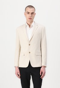 Beige textuurblazer met twee knopen, geknipt revers en voorzakken; gecombineerd met een wit overhemd en zwarte broek.