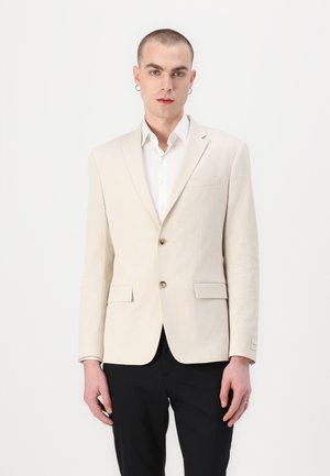 Beige textured blazer with two buttons, notched lapels, and front pockets; paired with a white shirt and black trousers.