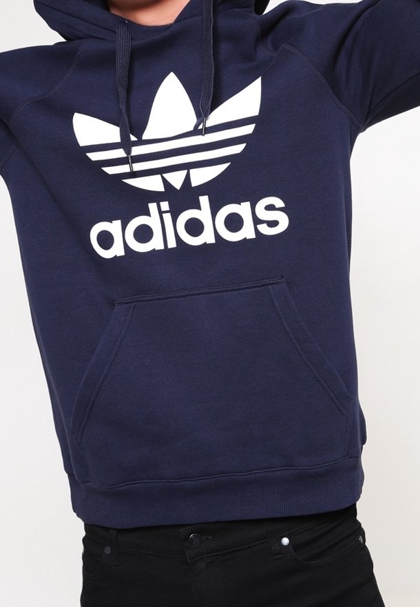 Námořnická modrá mikina s kapucí, která má velké bílé logo Adidas a tři bílé pruhy na hrudi. Obsahuje klokaní kapsu.