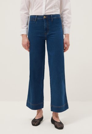 Femme portant un jean large bleu foncé avec une chemise blanche et des chaussures plates noires tissées, debout devant un fond uni.