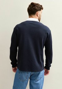 Mörkblå långärmad polo med vit krage, tillverkad av mjukt material, har en avslappnad passform och minimal sömnadsdetalj på baksidan.