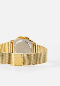 Montre avec un bracelet en maille dorée, boîtier rectangulaire, surface texturée et détails de fermoir. L'arrière présente une surface métallique argentée lisse.