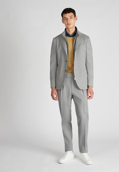 Giovane uomo con un completo grigio chiaro, maglione senape, camicia teal e sneakers bianche, in piedi davanti a uno sfondo chiaro e neutro.