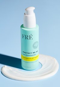 Bottiglia cilindrica blu chiaro con pompa bianca, etichettata "FRÉ Protect Me 50", accanto a un vortice di crema solare bianca su uno sfondo blu.