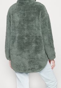 Polarowy sweter w stonowanej zieleni, z wysokim kołnierzem i długimi rękawami. Miękka, teksturowana tkanina z zaokrąglonym dołem z tyłu.