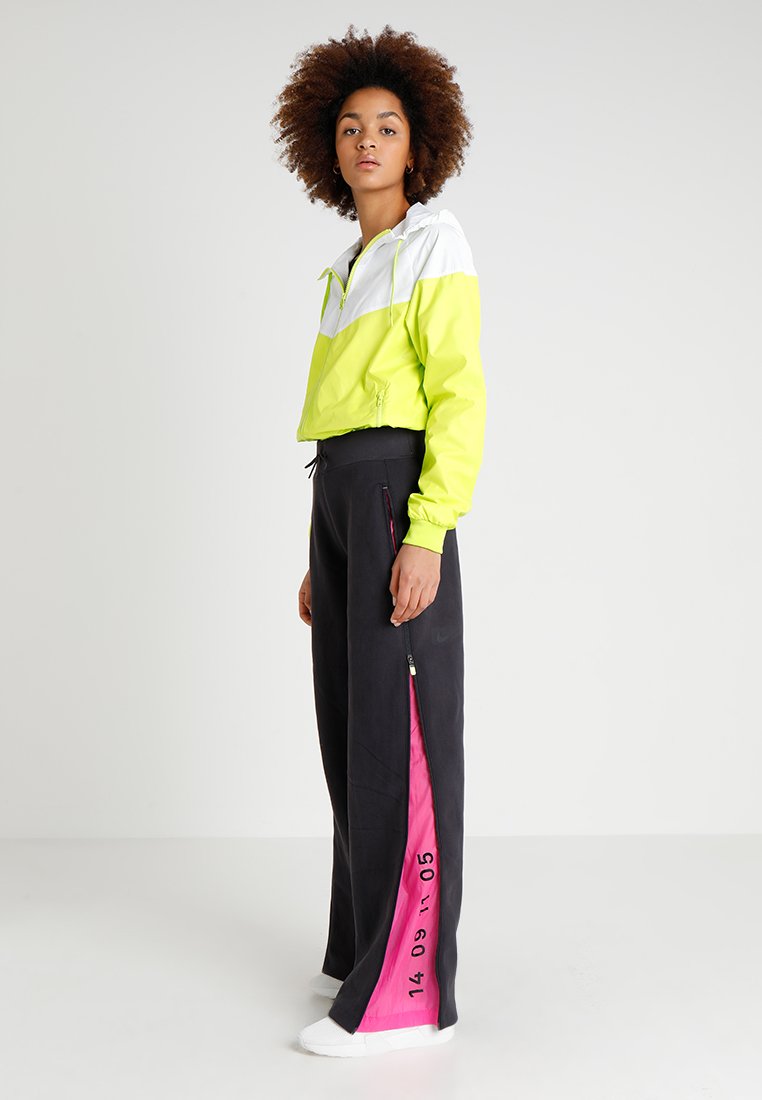 Veste coupe-vent de couleur jaune vif et blanche, associée à un pantalon noir avec des zippers latéraux roses et des chiffres imprimés. Chaussures blanches.