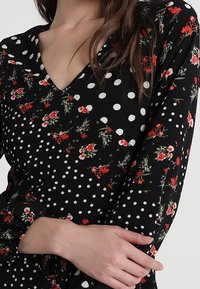 Tessuto nero con pois bianchi e motivo floreale rosso. Design con scollo a V, maniche lunghe e dettaglio arricciato al polso.