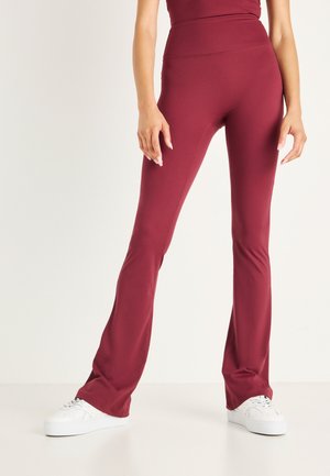 Femme portant des leggings évasés bordeaux taille haute et des baskets blanches, debout devant un fond clair uni.