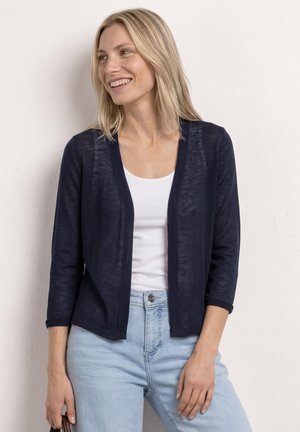 Uśmiechnięta blondynka, ubrana w granatowy kardigan na biały top i jasnoniebieskie dżinsy, stojąca na tle jednolitej białej ściany.