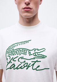 Νεαρός φορώντας λευκό μπλουζάκι με μεγάλο πράσινο κροκόδειλο και στυλιζαρισμένο κείμενο "Lacoste" μπροστά.