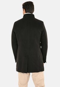 Cappotto di lana nero con collo alto, spacco posteriore e silhouette su misura. Tessuto uniforme senza motivi visibili o dettagli hardware.