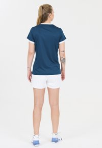 Urheilullinen teal-värinen urheilupaita valkoisilla yksityiskohdilla, yhdistetty valkoisiin shortseihin. Sileä kangas, rento leikkaus ja näkyviä tatuointeja käsivarressa.