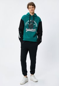 Sudadera color bloqueada en teal y negro con estampado gráfico y cordones, combinada con chándales negros. Zapatillas blancas completan el look.