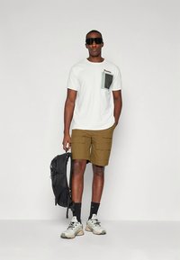 Hvit T-skjorte med korte ermer og grønn lomme, khakifargede cargoshorts, svart ryggsekk og beige treningssko med grå detaljer.