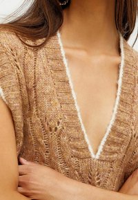 Strikket sweater i varm beige med dyb V-hals, der har et tekstureret mønster og hvid kant langs kanten. Korte ærmer.