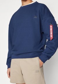 Felpa blu navy con scollo a girocollo, polsini a coste e tasca laterale con etichetta rossa. Indossata con pantaloni corti beige che presentano una finitura testurizzata.