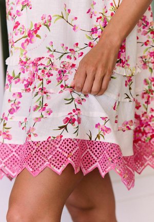 Hand hält weißes Kleid mit rosa Blumenmuster und gestufter rosa Spitzenkante vor Hintergrund mit bloßem Bein.