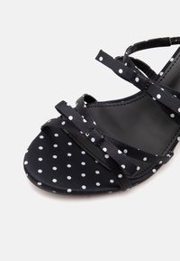 Sandale noire à pois blancs, avec des fines lanières et un petit nœud sur le devant au-dessus d’un orteil ouvert.