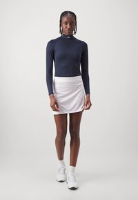 Tamsiai mėlynas ilgomis rankovėmis sportinis viršus su aukštu kaklu, derinamas su balta skortele, kurioje yra subtilus raštas, dėvima su baltais sportiniais bateliais.