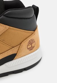 Gros plan sur le talon d'une basket beige et noire avec col rembourré, semelle blanche et logo arbre Timberland sur un panneau en cuir beige.