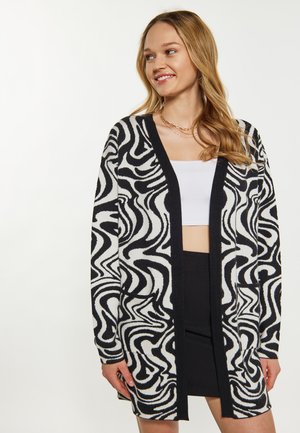Mladá žena se usmívá, má na sobě černo-bílý kardigan s vlnitým vzorem přes bílý crop top a černou sukní, na jednobarevném pozadí.