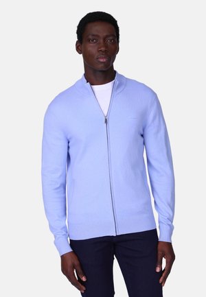 Homme portant un pull à fermeture éclair bleu clair sur une chemise blanche et un pantalon foncé, debout devant un fond uni.