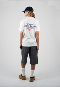 Γυναίκα φοράει λευκό φαρδύ T-shirt με το κείμενο «Απαγορευμένοι σύγχρονοι yuppies», κοντά ριγέ σορτς μέχρι το γόνατο, καφέ παπούτσια, άσπρες κάλτσες και μαύρο καπέλο.