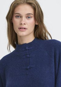 Tumši zila džemperis ar augstu kaklu, izgatavots no mīksta, teksturēta auduma. Iezīmē dekoratīvas pogas un ribotas daļas priekšā.