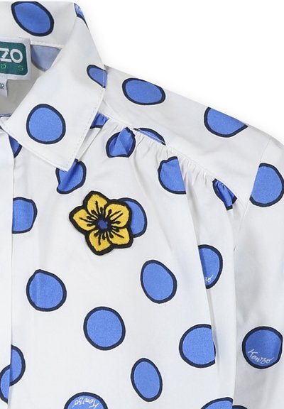 Chemise blanche avec de grands pois bleus, présentant une fleur brodée jaune à l'avant. Col standard et détails froncés.