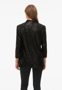 Zwarte blazer met pailletten, voorzien van een kraag en driekwartmouwen, met een gestructureerd oppervlak dat licht weerkaatst. Draagt over een strakke broek.