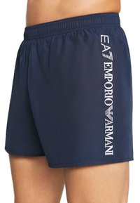 Bañador azul marino con cintura elástica, que presenta un logotipo blanco de "Emporio Armani" impreso verticalmente en el lado izquierdo. Textura suave.