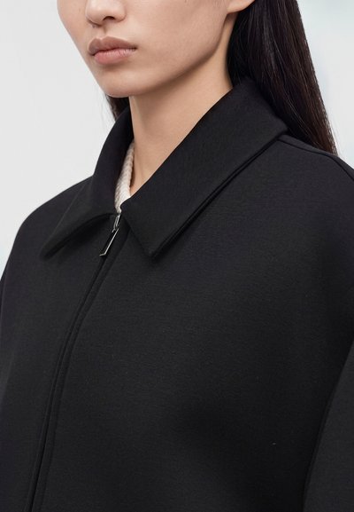 Gros plan d'une personne portant une veste noire zippée avec un large col et un vêtement intérieur blanc texturé visible au niveau de l'encolure.
