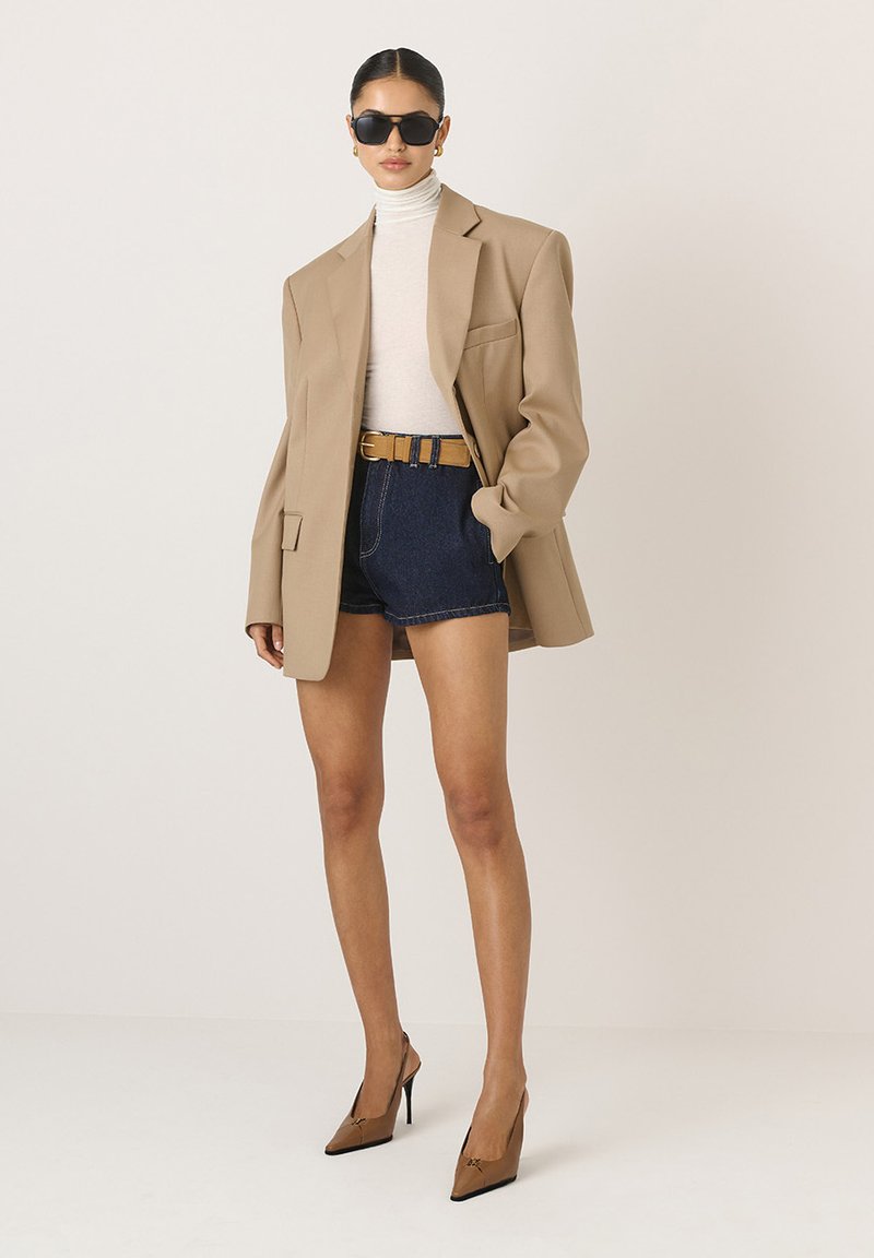 Beige oversized blézer egy hozzád simuló krémszínű garbó felett, sötét farmer rövidnadrággal és barna magassarkú cipővel párosítva, napszemüveggel kiegészítve.