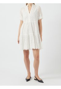 Kvinna som bär en vit ögla spetskortärmad klänning med volangkjol och svarta slingbacks med spetsig tå mot en vit bakgrund.