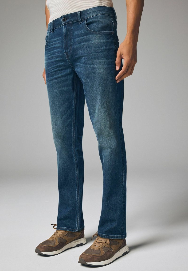 Donkerblauwe denim jeans met een rechte pijp, lichte vervaging en vijf-pocket stijl, gedragen met bruine en beige sportschoenen.