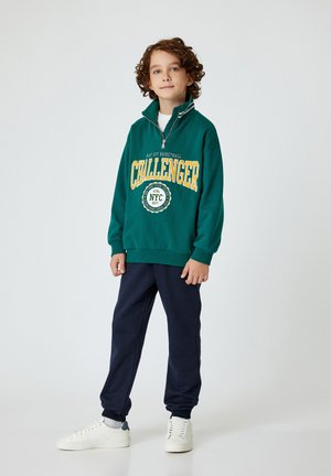 Vihreä fleece-sweatshirt, jossa on keltainen "CHALLENGER" -teksti ja NYC-logo; yhdistettynä tummansinisiin housuihin ja valkoisiin lenkkitossuihin; neljännesvetoketjukaulus.