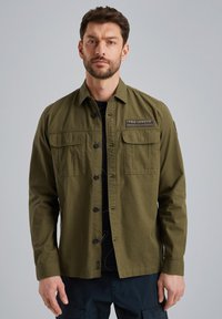 Camicia button-up verde oliva con due tasche sul petto, una patch tessuta etichettata "PME Legend" e maniche lunghe. Il tessuto ha una finitura testurizzata.