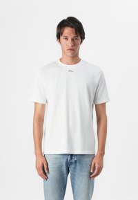Tricou alb din bumbac, cu mâneci scurte, guler rotund, accent logo subtil lângă guler, asortat cu blugi din denim de culoare albastră deschisă.