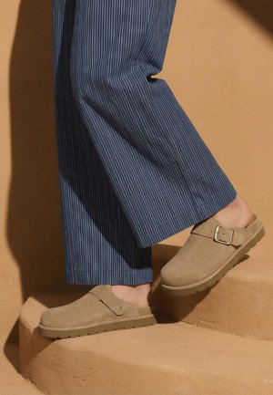 Picioare încălțate cu saboți slip-on bej cu o cataramă, călcând pe scări bej, asortați cu pantaloni largi albaștri cu dungi subțiri.